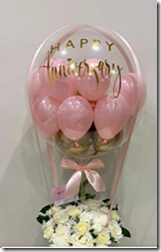 Pink balloons stuffed in a transparent air filled balloon with Print Happy BirthDay tied to basket of 12 white roses